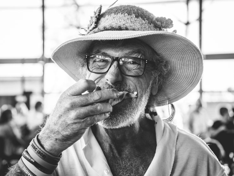 Image of An older man with glasses and a wide-brimmed hat decorated with a flower eats whilst looking at the camera. He wears several wristbands and a collared shirt. The background is blurred with people and large windows.