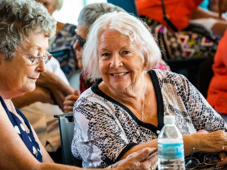 Image of Two older women sitting together indoors, one smiles at the camera while the other looks down. They are surrounded by people and there is a water bottle on the table in front of them.
