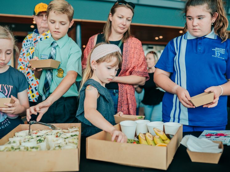 Image of A group of children and a woman stand at a table selecting food from trays filled with sandwiches, fruit, and cups, during a casual indoor event.