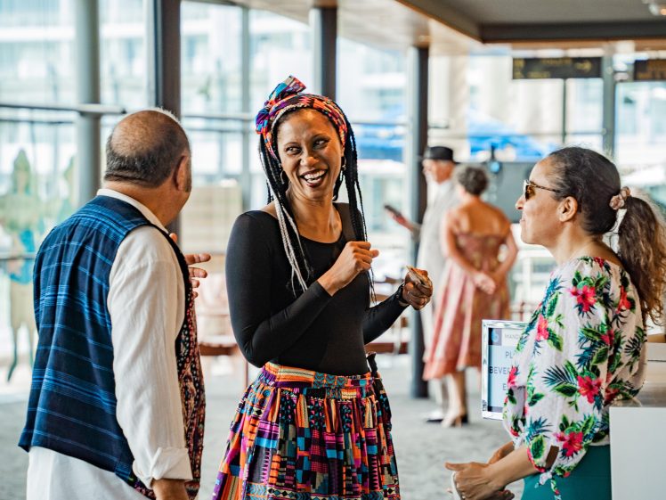 Image of Three people in colourful, patterned clothing are smiling and talking indoors near large windows. One woman wears a vibrant headwrap and skirt, while the other two, a man and a woman, are warmly engaged in conversation.