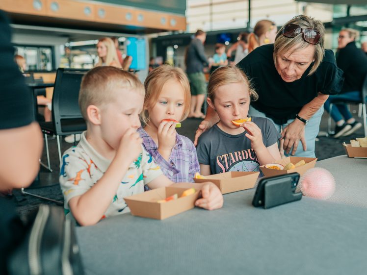 Image of Three children sitting at a table eat snacks from cardboard trays while watching something on a mobile phone. An adult woman leans over to look at the screen with them. Other people are visible in the background.