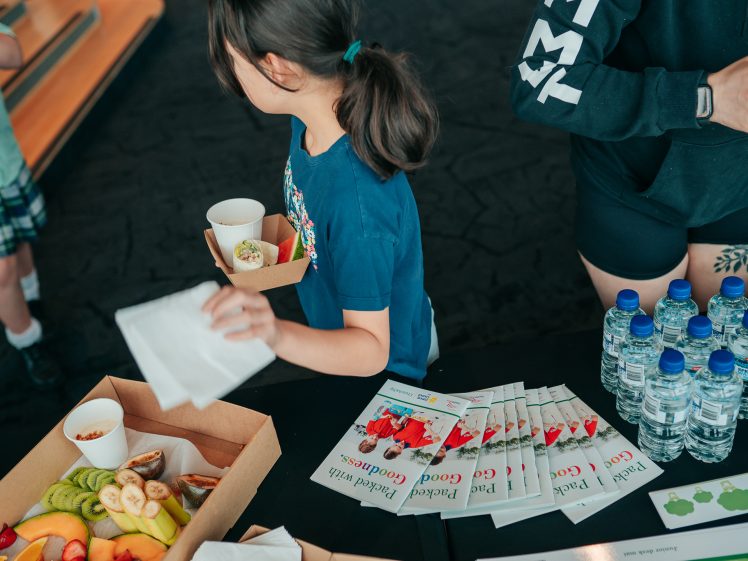 Image of A child holds a drink and a food tray near a table with water bottles, brochures, and trays of sliced fruit. Another person is seen beside the child, partially out of shot.