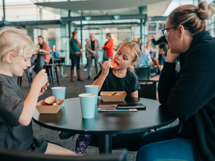 Image of Two young children and an adult sit at a round table in a bright canteen, eating food from cardboard trays and drinking from blue cups. Other people and large windows are visible in the background.