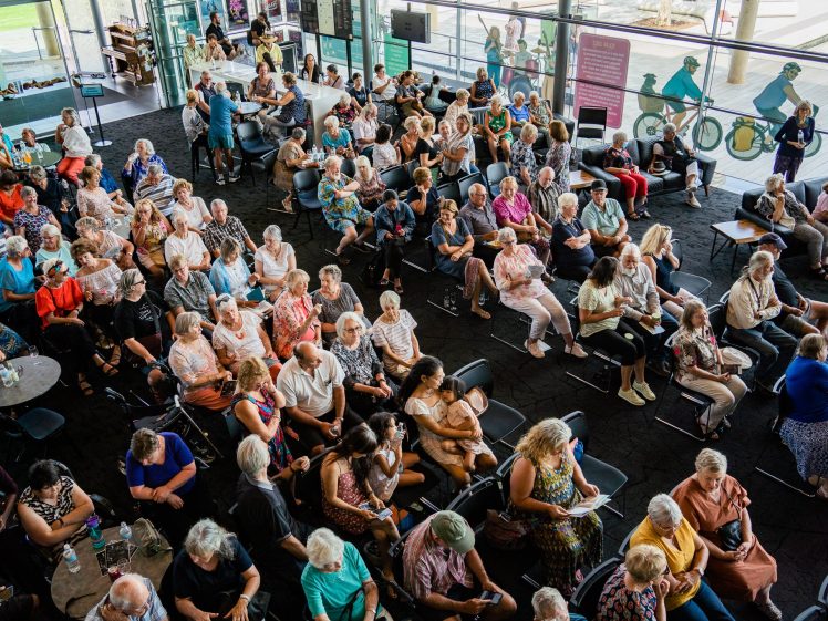 Image of A large group of people, mostly older adults, sit closely together in rows of chairs at an indoor event space with large windows, chatting and waiting for an event to begin.