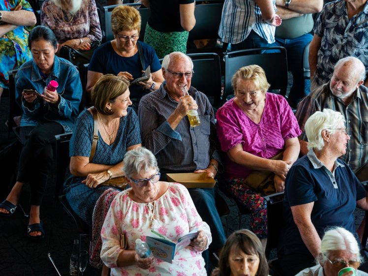 Image of A group of older adults sit closely together in rows of chairs, chatting, laughing, and looking at programmes or phones. The scene appears lively and social, with various expressions and interactions among the group.