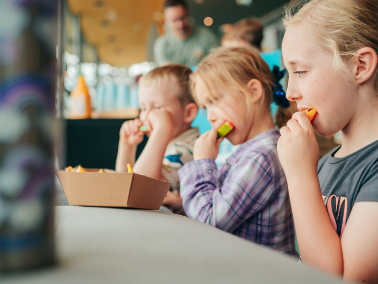 Image of Three children sit in a row indoors, eating slices of fruit or vegetables from a cardboard carton. The focus is on the children while the background is softly blurred.