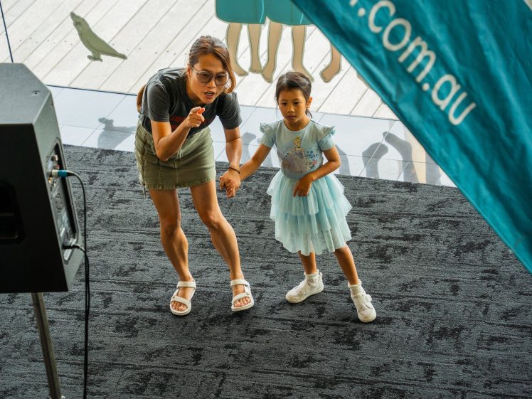 Image of A woman and a young girl in a blue dress stand on a dark carpeted floor, holding hands and looking intently ahead. The woman is pointing, and a teal banner is visible in the foreground.