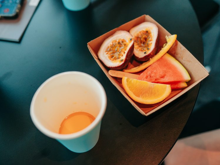 Image of A paper cup with some orange liquid sits on a black table next to a cardboard tray holding passion fruit halves, a slice of watermelon, and a slice of orange.