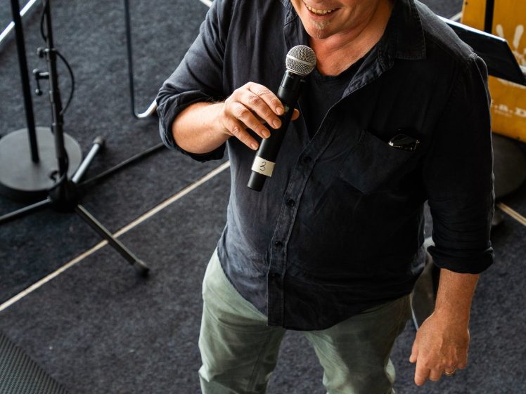 Image of A man with grey hair and glasses smiles whilst holding a microphone, standing on a dark carpeted floor near a chair, microphone stand, and wooden box. He is wearing a dark shirt and grey trousers.