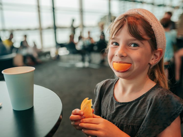Image of A young girl with a playful expression holds an orange segment in her hands and has another orange segment in her mouth, mimicking a big smile. She sits at a table with a paper cup in a busy indoor setting.