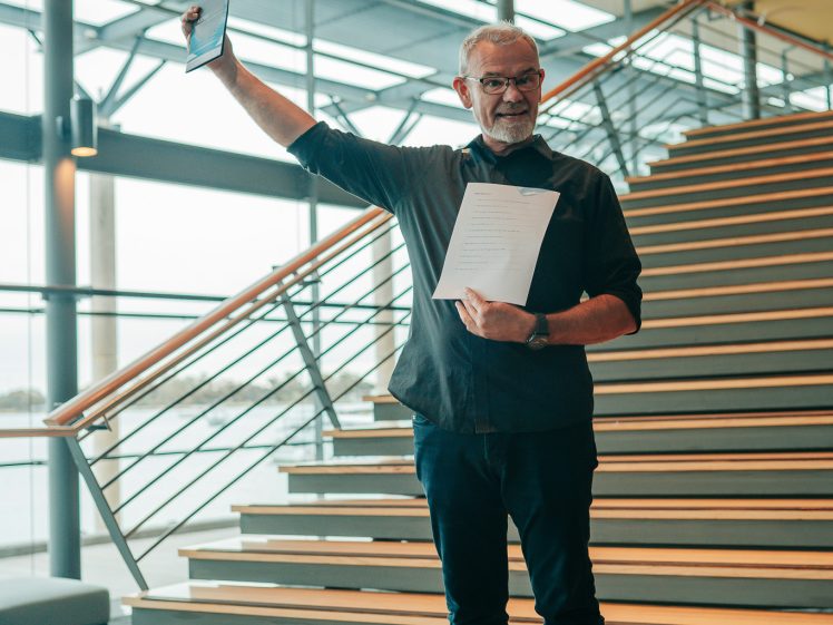 Image of A man with glasses stands on indoor stairs, smiling and holding a tablet up in one hand and papers in the other, in a modern building with large windows.