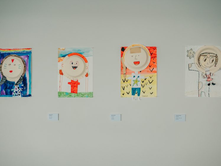 Image of Four colourful children's artworks are displayed on a wall. Each piece features a face made from a paper plate, decorated with various materials and drawings, and includes a mix of painted and drawn backgrounds.