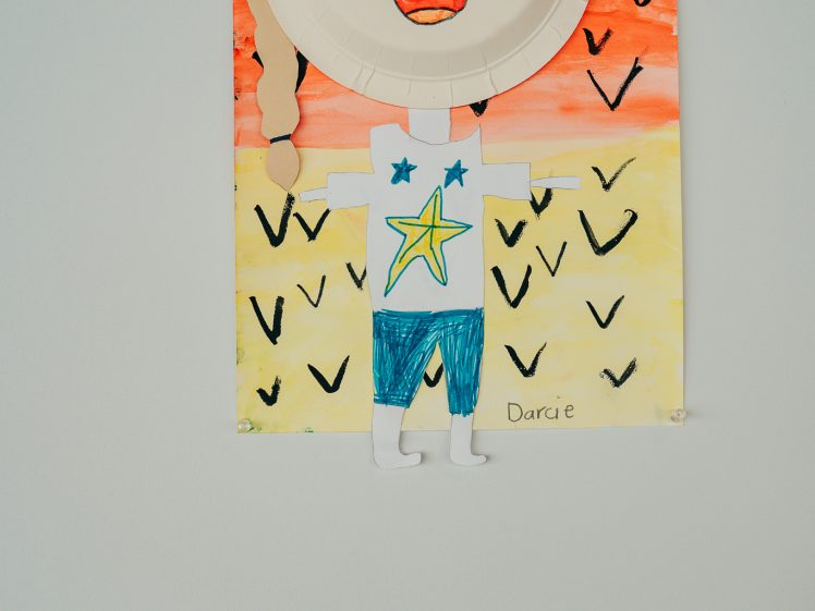 Image of A children's artwork features a smiling person made from a paper plate face, paper arms and legs, and a painted background with V shapes. The name Darcie is written on the bottom right. A small label is displayed on the wall nearby.