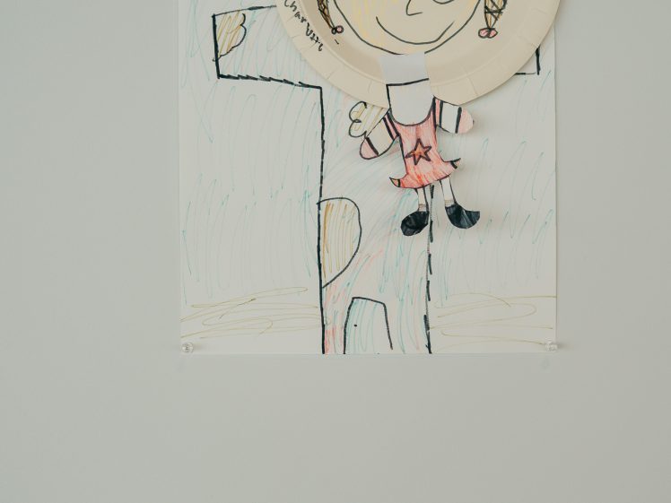 Image of A child’s drawing of a person with a paper plate for a face is pinned to a wall, featuring colourful crayon details and a sun in the corner. A small label is displayed below the artwork.