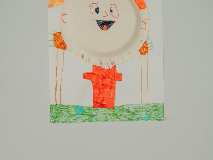 Image of Children's drawing of a person with a paper plate for a face, orange hair, and body, smiling, with arms outstretched, displayed on a wall with a small label beneath it.