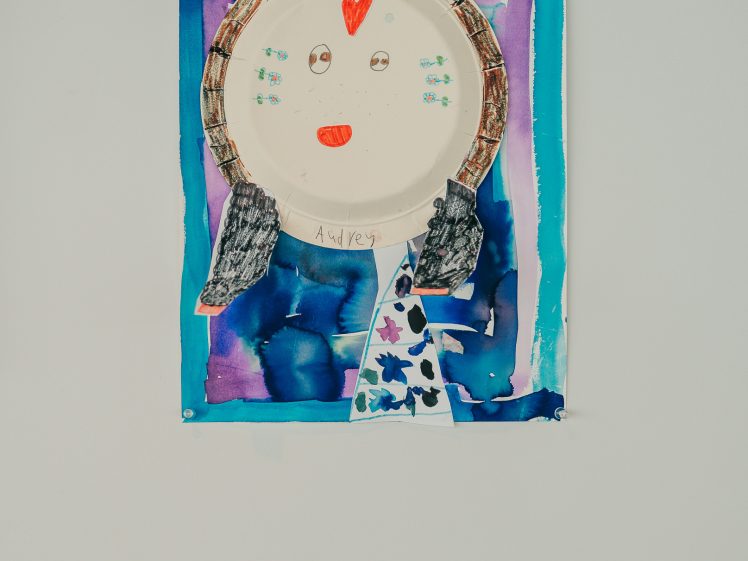 Image of A colourful, child-made portrait features a paper plate face with a red heart on the forehead, red mouth, and wool hair, set against a painted blue-purple background with a rainbow and abstract shapes. A label is displayed below.