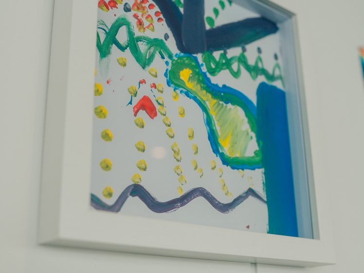 Image of A framed abstract painting features colourful shapes, dots, and wavy lines in blue, green, yellow, and red on a white background, displayed on a white wall.