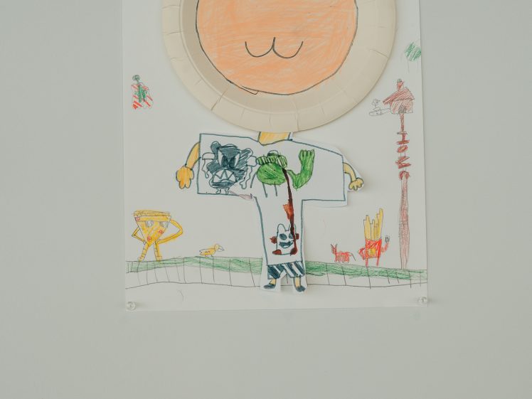 Image of Children’s drawing of a smiling person with a paper plate for a face, colourful body and arms, and playful background including trees, animals, and outdoor scenery, displayed on a wall with a small label below.