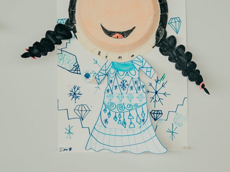 Image of A child’s drawing of a smiling queen with plaited hair, a blue crown, and a decorated dress, surrounded by blue snowflakes and diamonds. The artwork is displayed on a wall with a small label beneath it.