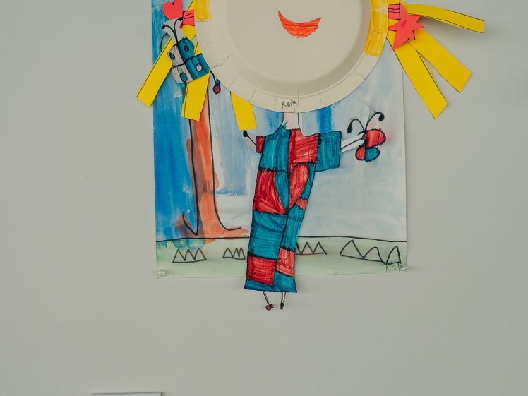 Image of A colourful children's art project depicting a smiling sun made from a paper plate, with yellow paper rays, a red-and-blue body, and a hand-drawn background featuring trees, grass, and butterflies.