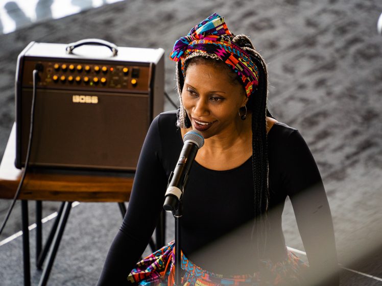 Image of A woman with plaited hair and a colourful headwrap sings or speaks into a microphone. She is wearing a black top and a patterned skirt, with a BOSS amplifier on a table behind her. The background is dark and blurred.