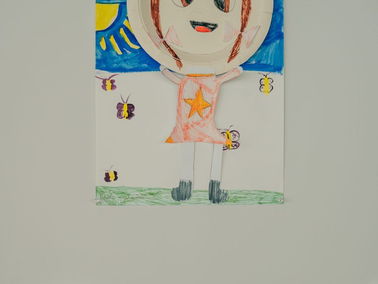 Image of A colourful child’s drawing of a smiling figure with a paper plate for a head, standing on grass with butterflies, under a blue sky with a bright yellow sun. The figure has brown hair, pointed ears, and a star on its dress.