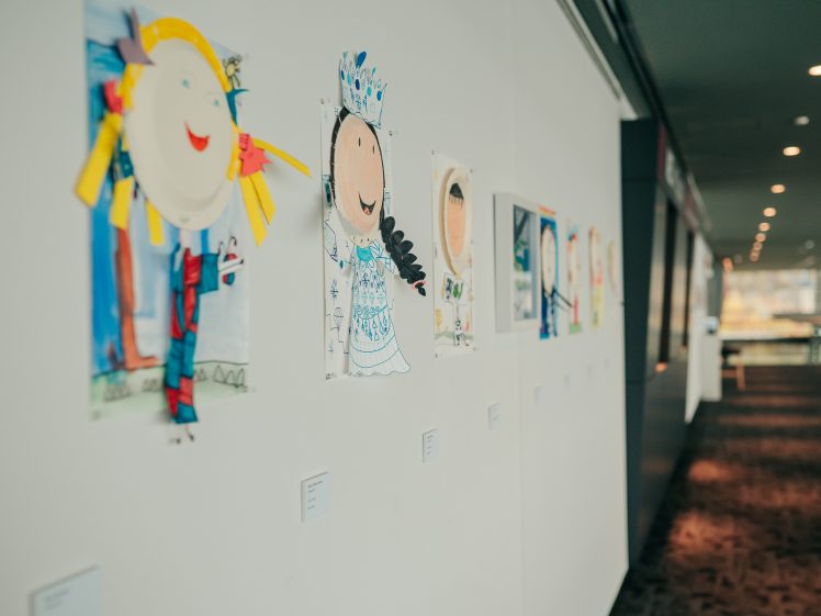 Image of Children’s artwork made from paper plates and colourful materials is displayed in a row on a white wall in a brightly lit corridor or gallery space. Each piece features a smiling face and unique decorations.