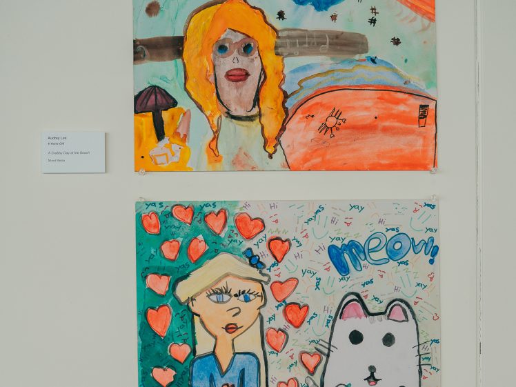 Image of Two colourful children's drawings on a wall. The top shows a person with orange hair, a sun, and bright colours. The bottom features a girl with blonde hair, many red hearts, and a white cat with meow and Pusheen written above.