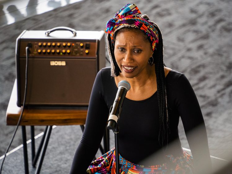 Image of A woman wearing a colourful headwrap and patterned skirt sings into a microphone. An amplifier sits on a small table behind her. She appears expressive and focused, performing indoors on a dark carpet.
