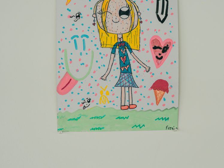 Image of A colourful, childlike drawing of a girl with blonde hair on a grassy patch, surrounded by playful symbols including a star, heart, ice cream, and smiley face, with a white wall and a small label beneath the artwork.