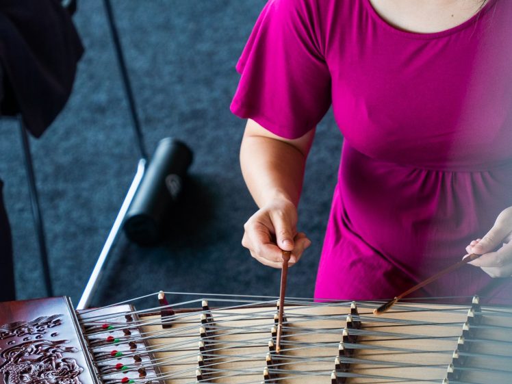 Image of A woman in a magenta dress plays a traditional string instrument, possibly a yangqin, using thin wooden beaters. She is seated, and the instrument has intricate carvings and colourful tuning pegs.