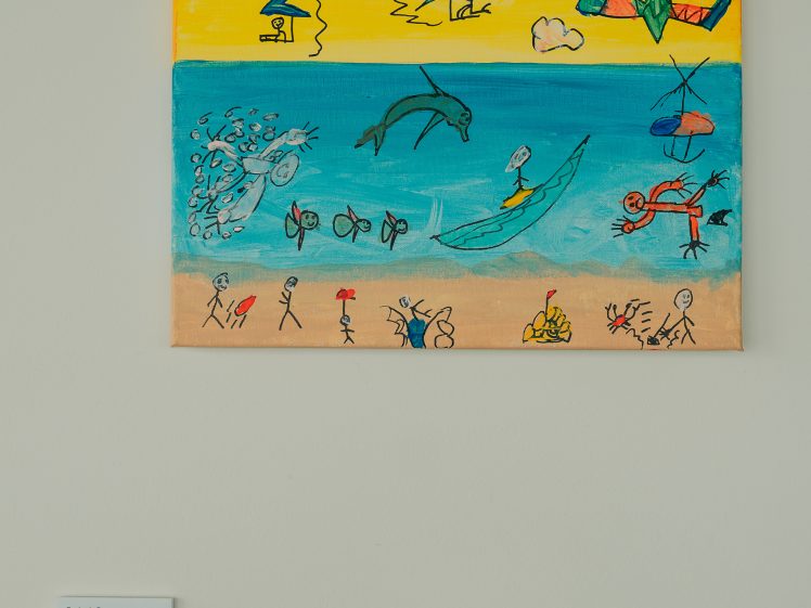 Image of A colourful drawing of a beach scene with stick figures, sea creatures, and people surfing or flying kites under a bright yellow sky. The artwork is displayed on a white wall with a small label below it.