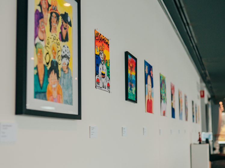 Image of Framed colourful drawings and paintings are displayed in a row on a white gallery wall, each with a small label beneath it, creating a vibrant art exhibition in a well-lit, modern space.