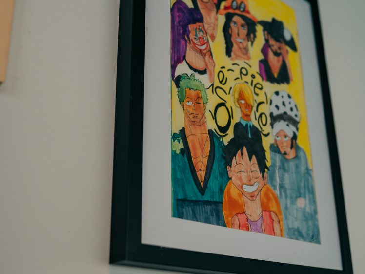 Image of Framed, colourful fan art of characters from the anime One Piece hangs on a white wall. A small label with text is positioned just below and to the left of the artwork.