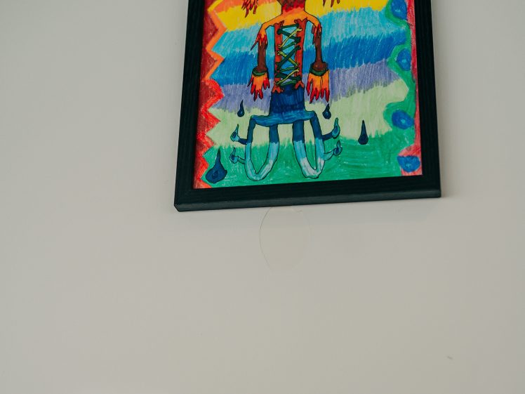 Image of Colourful childlike drawing in a black frame shows a smiling figure with red hair, blue clothing, and large shoes, set against a rainbow-striped background with zigzag and wavy patterns. A label is mounted below the artwork.