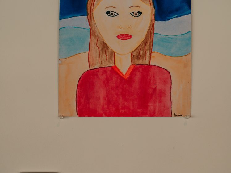 Image of A colourful painting of a girl with brown hair and a red shirt, set against a beach background with blue waves, a turtle, and an orange starfish in the sky. The painting is displayed on a wall with a label beneath it.