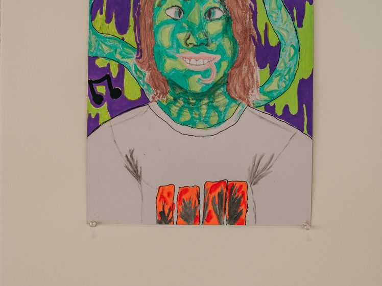 Image of A colourful drawing of a person with green skin and brown hair, with snakes behind their head and a purple and green background. The person is wearing a grey shirt with red marks on it. An art label is visible on the wall below.