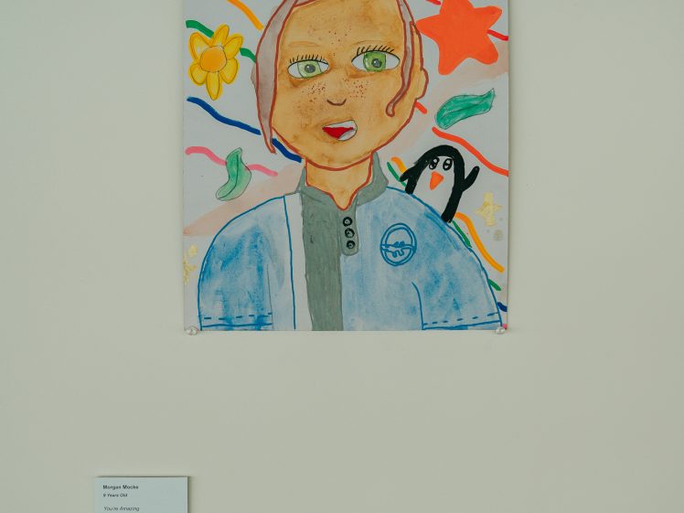 Image of A colourful drawing of a person with orange hair, surrounded by flowers, stars, and a penguin on their shoulder, displayed on a white wall with a small label below it.