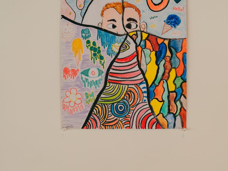 Image of A colourful, abstract drawing divided by black lines into sections with various patterns: faces, hearts, swirls, cartoon shapes, and bright splashes. The artwork hangs on a white wall above a small, labelled placard.
