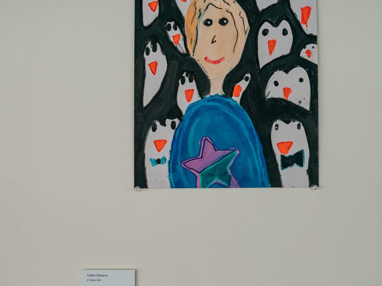 Image of A colourful painting of a person holding their head, surrounded by penguins with orange beaks, is displayed on a wall. A small placard with information about the artwork is mounted below it.