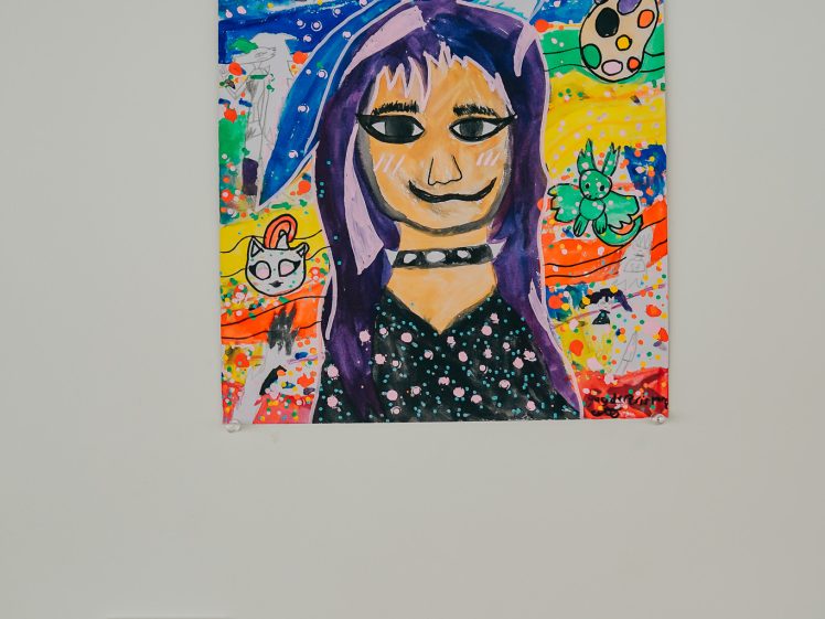 Image of Colourful painting of a person with purple hair and cat ears, surrounded by playful cat figures, a paw print, palette, and vibrant, abstract background. Artwork is displayed on a white wall with a label below.
