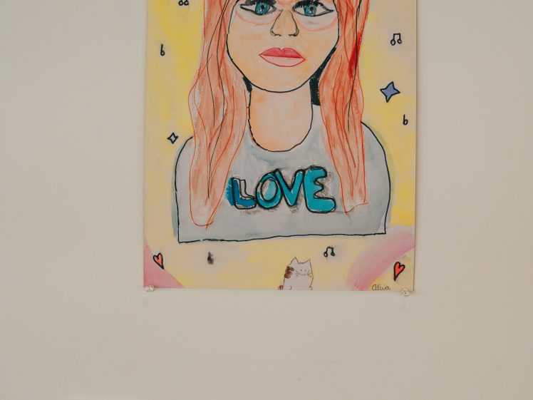 Image of A colourful drawing of a woman with long red hair, glasses, and a shirt that says LOVE. The background has hearts, stars, and musical notes. A small cat appears at the bottom centre of the artwork.