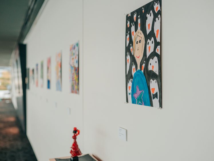 Image of A close-up of a colourful painting on a white gallery wall shows a person surrounded by cartoon penguins. Other artworks are visible in a row along the wall, and a small red sculpture sits on a nearby surface.