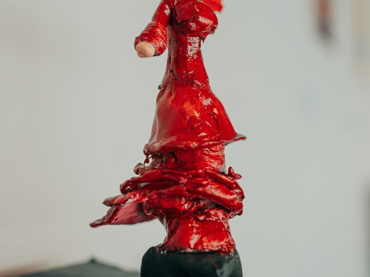 Image of A small clay sculpture of a person in a red dress with a flower in their hair stands on a black plinth against a blurred background.
