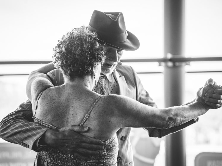 Image of An older couple dances closely together, the man wearing a hat and suit, and the woman in a sleeveless dress. They embrace while dancing, with soft natural light in the background. The photo is black and white.