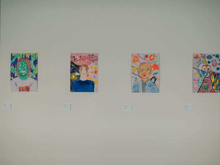 Image of Four colourful, abstract portrait paintings hang on a white wall, each with a small label below. The artworks feature different faces with vivid backgrounds and imaginative, vibrant designs.