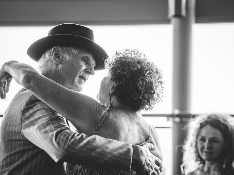 Image of An older couple dances closely, sharing a tender moment. The man wears a hat and a striped suit. A woman with curly hair stands in the background, watching them and smiling. The photo is in black and white.