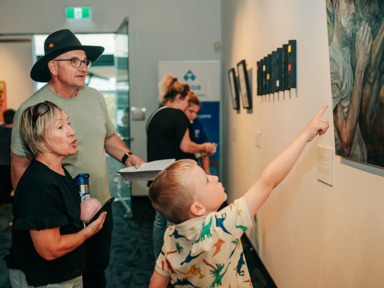 Image of A young boy in a dinosaur shirt excitedly points at artwork on a gallery wall, whilst two adults beside him watch and hold papers. Other visitors are seen in the background.
