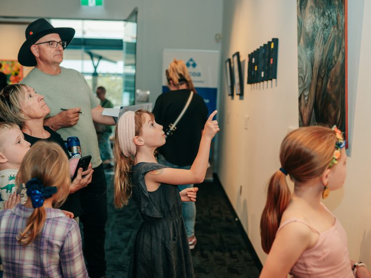 Image of A group of children and adults look at artwork displayed on a gallery wall. One girl in a grey dress points at a painting, while others observe with interest. The setting appears bright and modern.