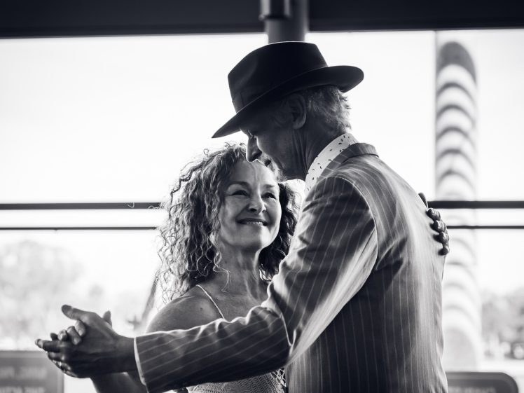 Image of An older couple dance closely together, smiling. The woman has curly hair, and the man wears a pinstripe suit and hat. They appear happy and content, sharing a tender moment indoors. The image is in black and white.